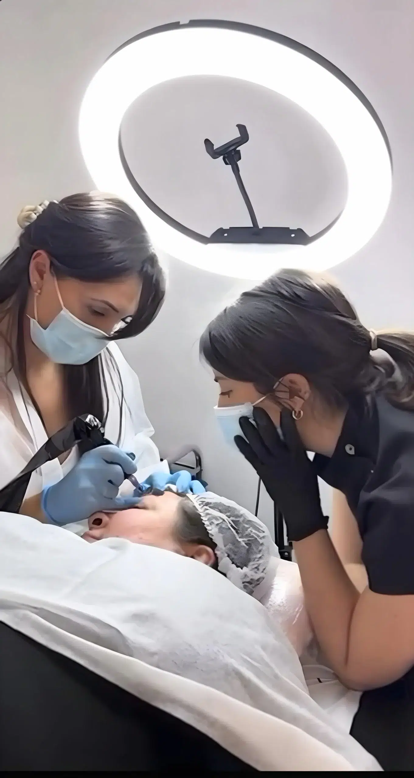 micropigmentacion Dos personas con mascarillas y guantes realizan un procedimiento de micropigmentación en una persona acostada. La persona de la izquierda utiliza un dispositivo portátil en el rostro de la clienta. Un anillo de luz ilumina la escena, y la clienta lleva una gorra. Monica Aranguez