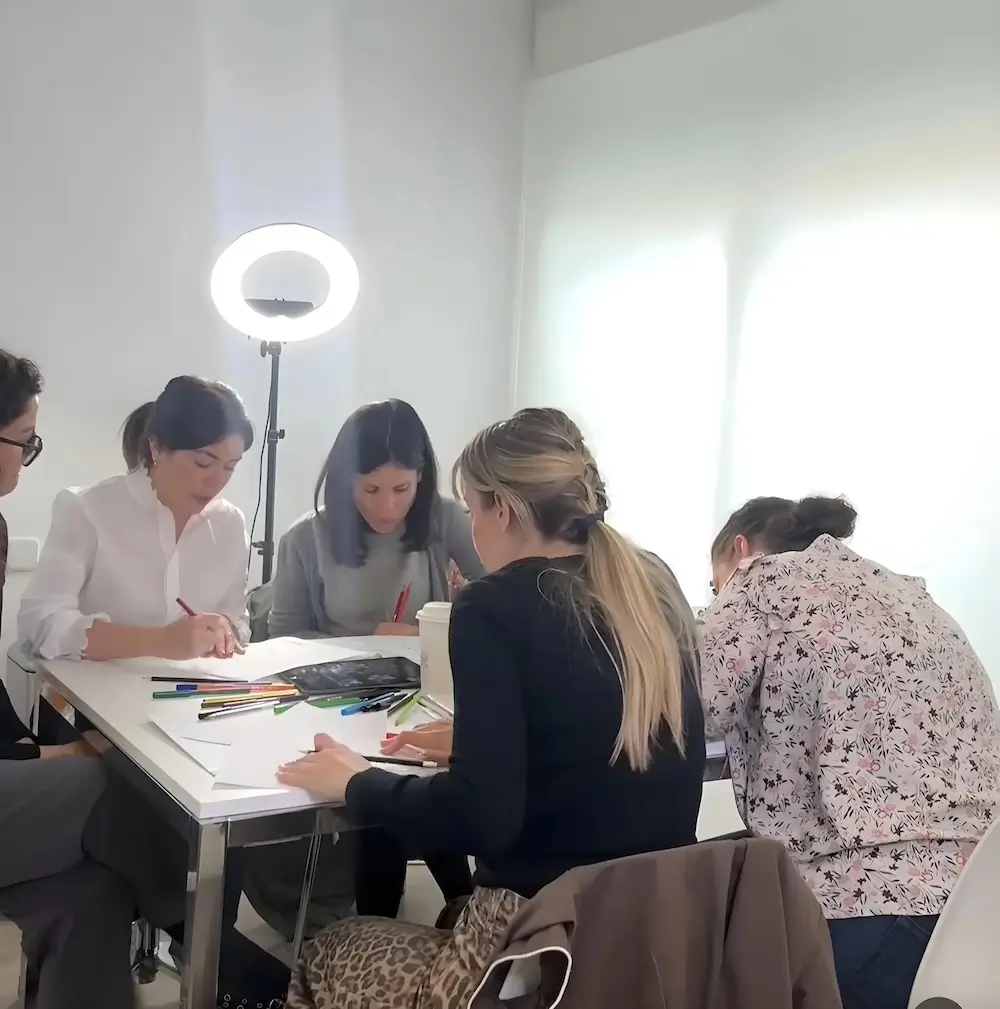 micropigmentacion Un grupo de personas se sienta alrededor de una mesa en una habitación iluminada, escribiendo o dibujando mientras un anillo de luz ilumina el espacio de trabajo. Parecen asistir a uno de esos cursos creativos grupales que dan vida a las ideas con cada pincelada. Monica Aranguez