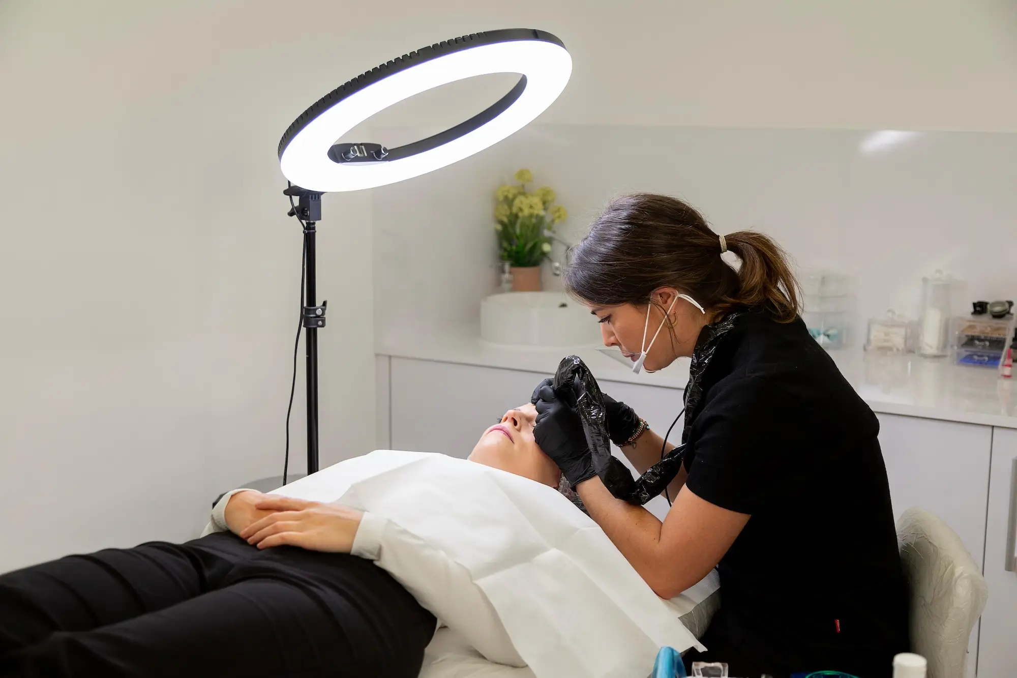 micropigmentacion Una cosmetóloga, con guantes y mascarilla, realiza meticulosamente una micropigmentación terapéutica en una persona recostada en un sillón. Un anillo de luz proyecta su luz sobre la habitación blanca e inmaculada. Monica Aranguez