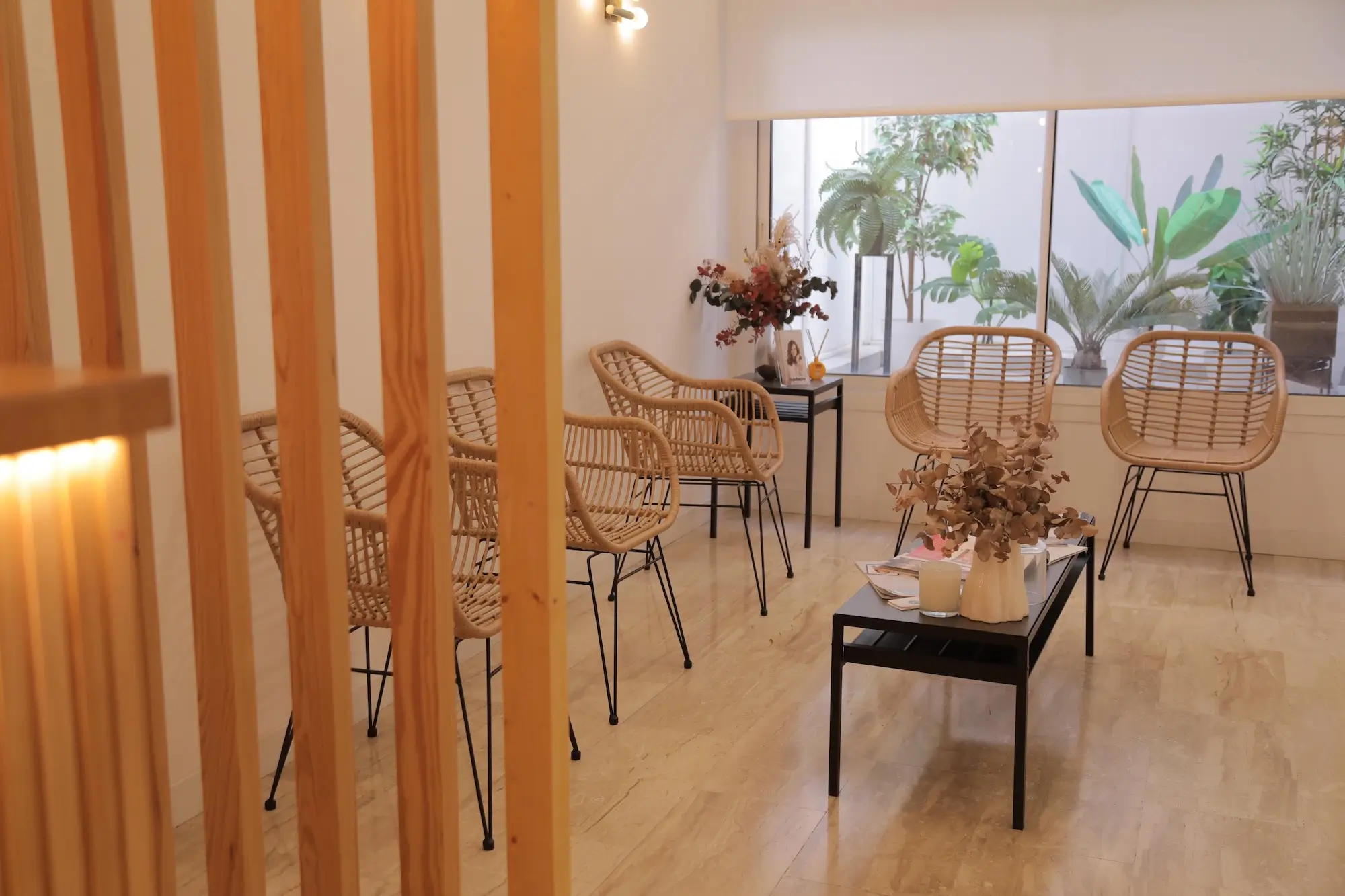 micropigmentacion Una moderna sala de espera con sillas de mimbre rodea una mesa de centro negra con revistas y un jarrón. Listones de madera dividen el espacio, dejando ver folletos de micropigmentación terapéutica a través de grandes ventanales. El suelo de madera clara complementa a la perfección las paredes blancas. Monica Aranguez