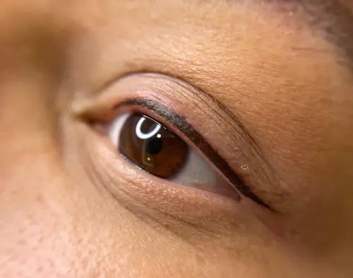 micropigmentacion Primer plano de un ojo humano con iris marrón y párpado visible. El párpado presenta delineador negro, posiblemente realzado mediante micropigmentación ocular. La piel luce suave y con textura natural. Monica Aranguez