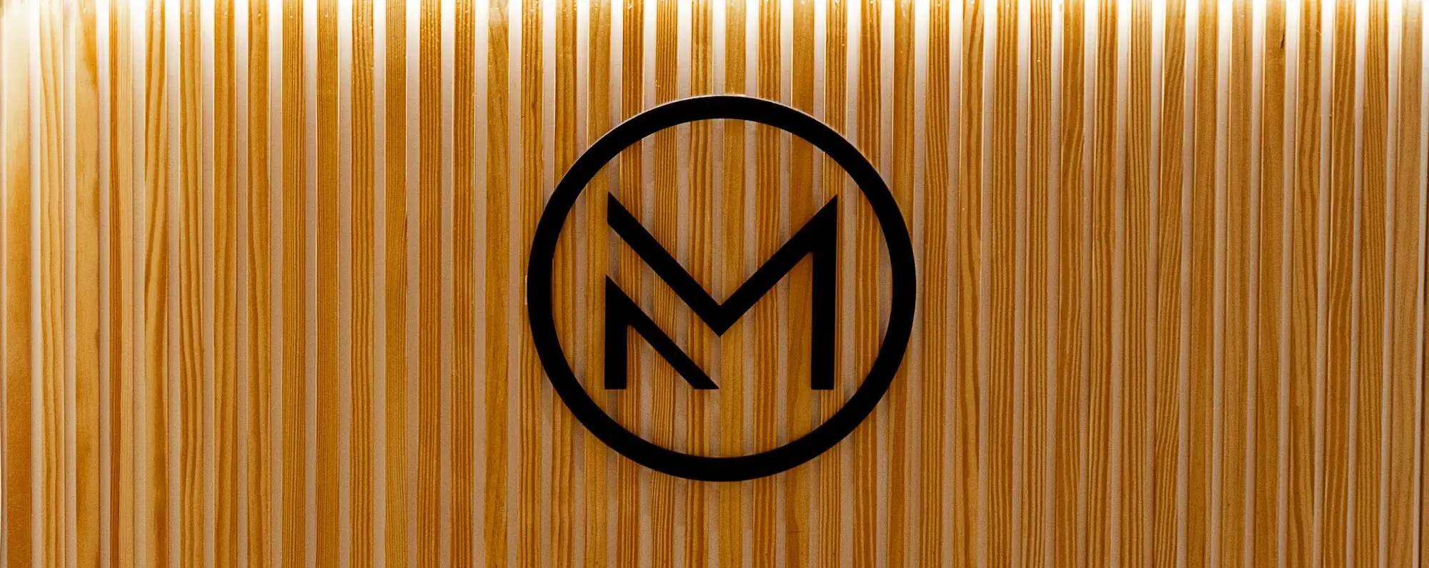 micropigmentacion Un círculo negro con la letra "M" en su interior está centrado sobre un fondo de madera texturizado con ranuras verticales. Monica Aranguez