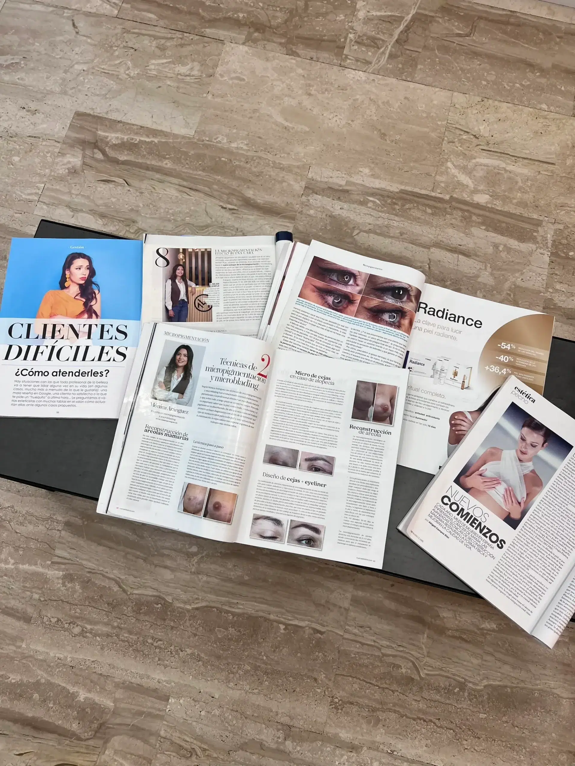 micropigmentacion Revistas de belleza y moda abiertas sobre una mesa negra. La portada dice "CLIENTES DIFÍCILES ¿Cómo atenderles?". Se ven varios artículos e imágenes sobre cuidado de la piel y cosmética, sobre un fondo de suelo de piedra. Monica Aranguez