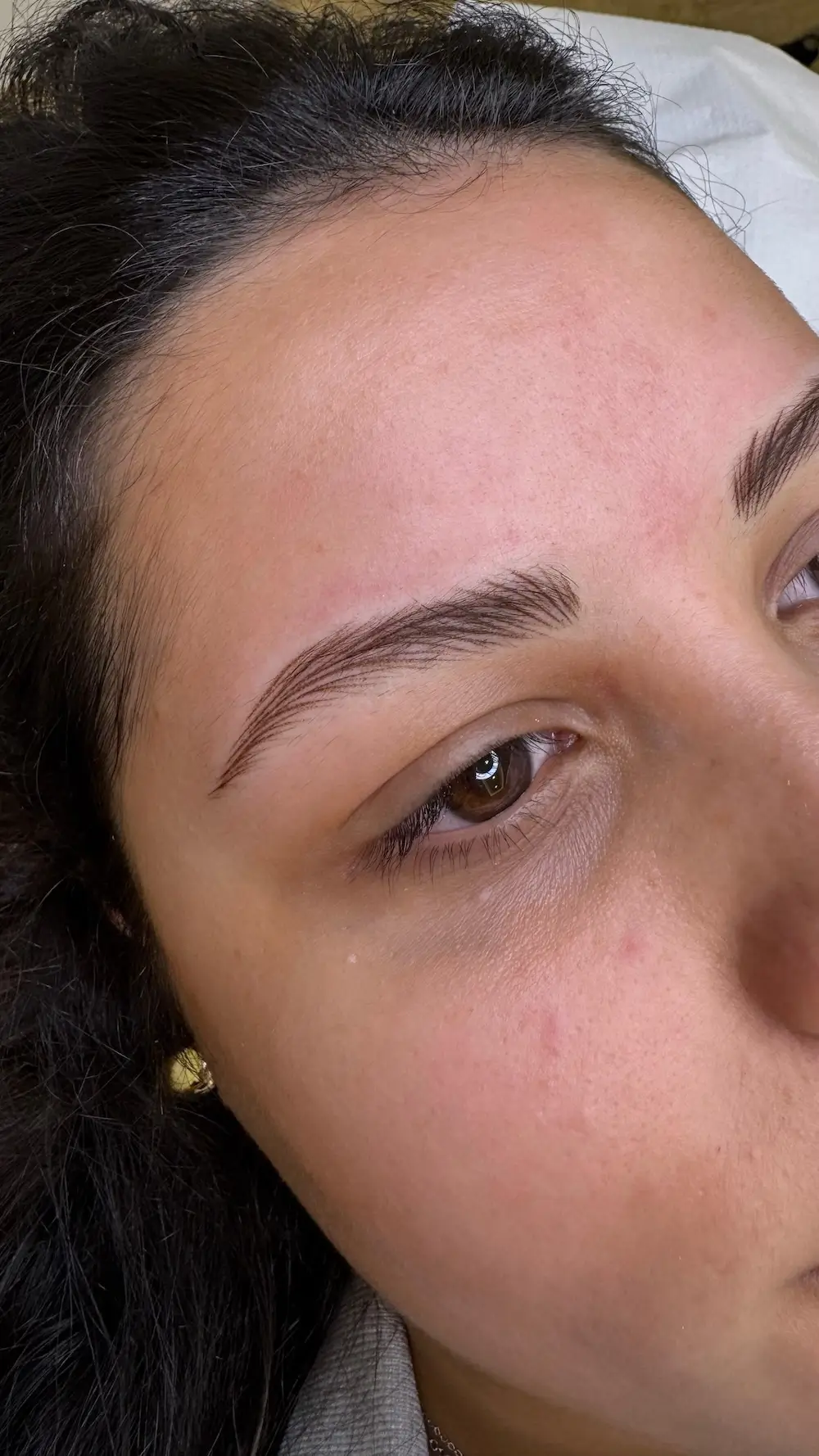 micropigmentacion Un primer plano del rostro de una persona muestra unas cejas recién depiladas con micropigmentación. La piel luce suave y las cejas, perfectamente delineadas, presentan arcos definidos. Con cabello oscuro, la persona está tumbada sobre una superficie blanca. Monica Aranguez
